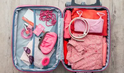 Open suitcase neatly organised with vibrant pink and red clothing, trendy accessories, sunglasses, and travel-size toiletries.