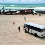 White coach parked on sandy Fraser Island beach by holidaymakers and iconic rusted shipwreck, K'gari Day Tour Ex Noosa, Australia.