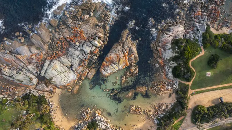 Stunning aerial view of vibrant orange-streaked rocky shoreline, pristine sandy beach, crystal-clear waters, and lush green countryside.