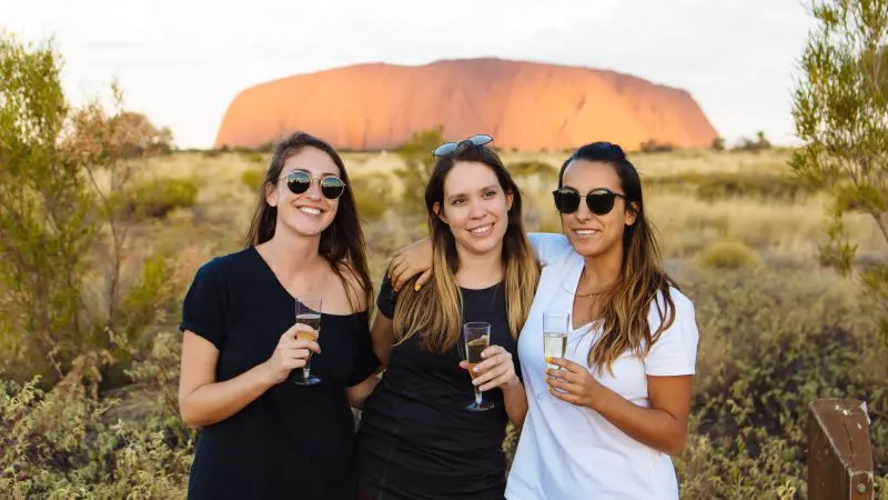 Three women enjoy drinks and smile in front of Uluru on a 3 Day Uluru Rock The Centre Tour from Alice Springs, capturing adventure.