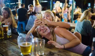 Two friends smiling and taking a selfie at a vibrant outdoor bar—book our 16-day travel deal: just £99/day for unforgettable fun.