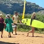 Four people walk along a sandy path, filming possibly for Celebtime Tours or 1 Day Home Away Tour, with a bright yellow surfboard nearby.