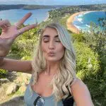 Smiling woman with long blonde hair takes a selfie by the sea, flashing a peace sign on her 1 Day Home Away Tour coastal adventure.