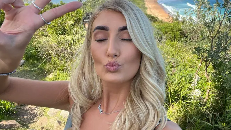 Smiling woman with long blonde hair takes a selfie by the sea, flashing a peace sign on her 1 Day Home Away Tour coastal adventure.