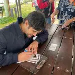 A celebrity signs autographs at a Celebtime Meet an Actor tour, sitting at a picnic table whilst fans eagerly wait in queue nearby.