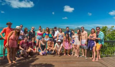 Travellers on the Original 21-day tour smile for a group photo on a wooden deck, framed by lush greenery and a vivid blue sky.
