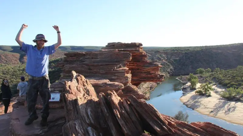 Traveller on rugged cliffs with arms raised, overlooking winding river and lush scenery during 8 Day Perth to Exmouth Return Tour adventure.