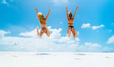 Two women in colourful bikinis leap with excitement on a bright beach, showcasing a top-value 16-day holiday package at £99 per day.
