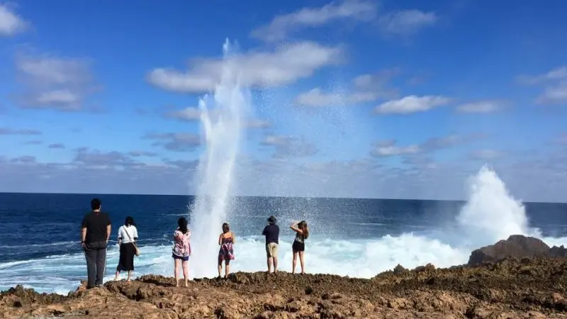 Group of six travellers on rocky coastline admire ocean waves during 8 Day Perth to Exmouth Return Tour beneath blue skies with clouds.