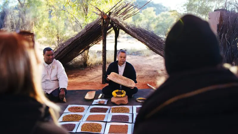 Two travellers showcase authentic native foods outdoors during a 3 Day Uluru Rock The Centre Tour from Alice Springs, Australia.