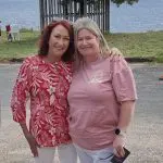 Two smiling women in red floral and pink tops by a serene lake during Celebtime Tours 1 Day Home Away Tour, enjoying scenic views.