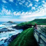 A couple enjoys scenic views from a wooden boardwalk of Phillip Island’s rugged coastline, lush green hills, and clear blue sky.