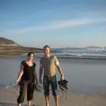 Two happy travellers stand barefoot on Wilsons Promontory’s sandy beach with Go West Tours, grinning beside scenic ocean waves.