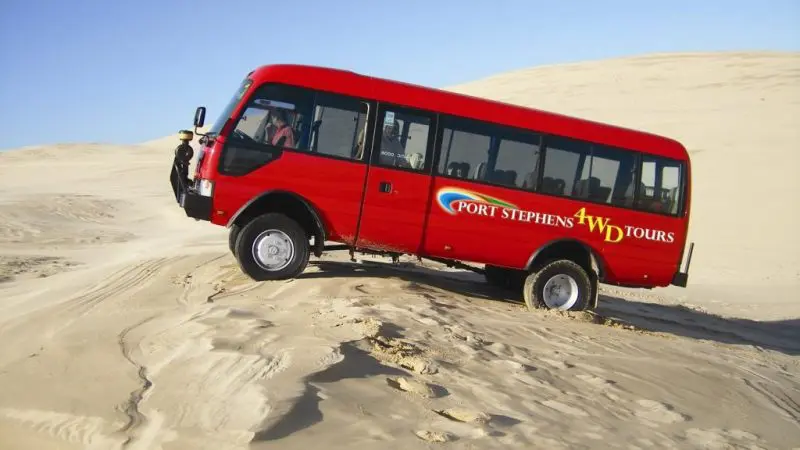 Experience the thrill as a red 1 Day Port Stephens 4WD tour coach ascends sandy dunes with Colourful Collective Travel adventure tours.