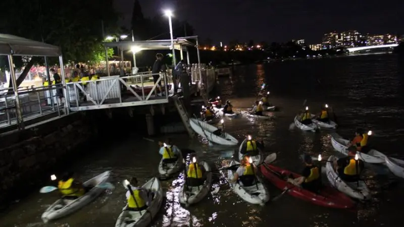 Kayakers in bright yellow vests with glowing lights paddle by a quay at night, illuminated city skyline and reflections in the background.