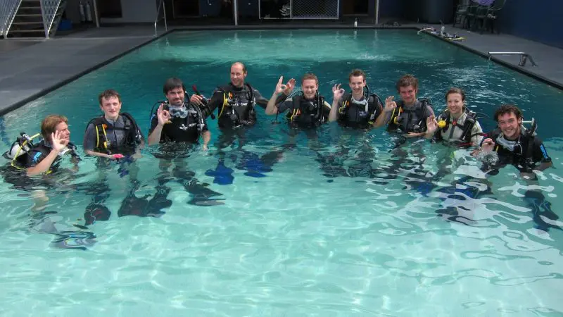 Ten scuba divers in full kit smile together in a swimming pool after completing a 3 Day 2 Night Open Water Dive Course for certification.