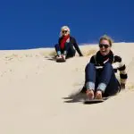 Two women sandboarding on golden dunes under the sun, smiling during a 1 Day Whale Watching Port Stephens adventure tour.
