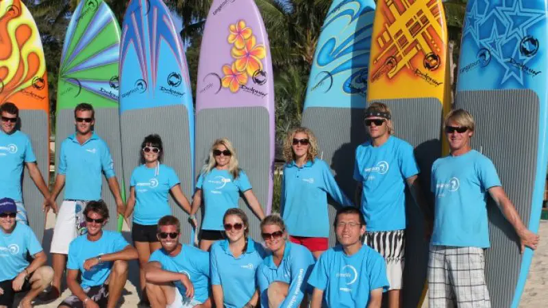 Participants in matching blue shirts stand on a sandy beach with paddleboards during the Adventure Moreton Island Wrecks Tour.