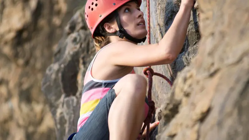 A female rock climber wearing a helmet and safety harness scales a challenging outdoor cliff during a guided climbing session.