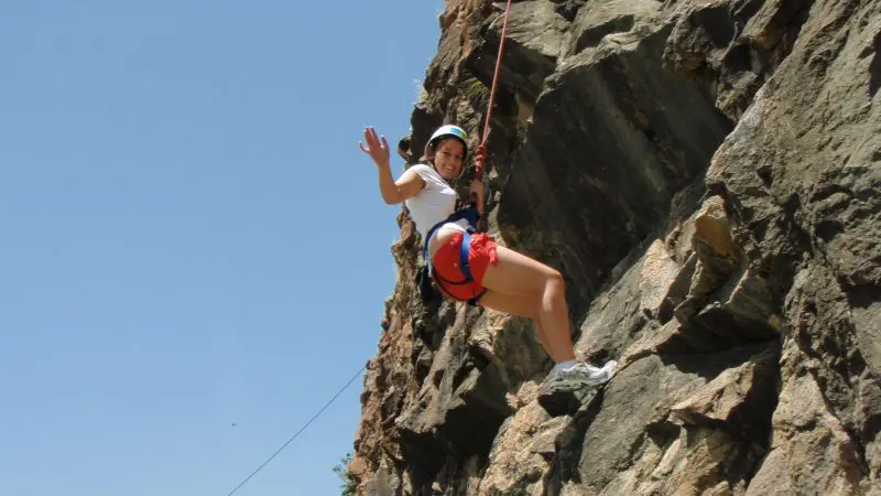 Adventurer smiles and waves during guided rock climbing session, scaling a rugged cliff face beneath clear blue sky, outdoor activity.