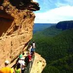 Adventurous hikers traverse a narrow cliffside trail on a 1 Day Blue Mountains Wildlife Park Tour, with breathtaking valley views.