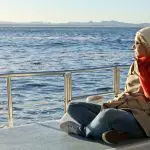Person in coat and red scarf aboard 1 Day Whale Watching Port Stephens boat, looking at scenic mountains under clear blue sky.