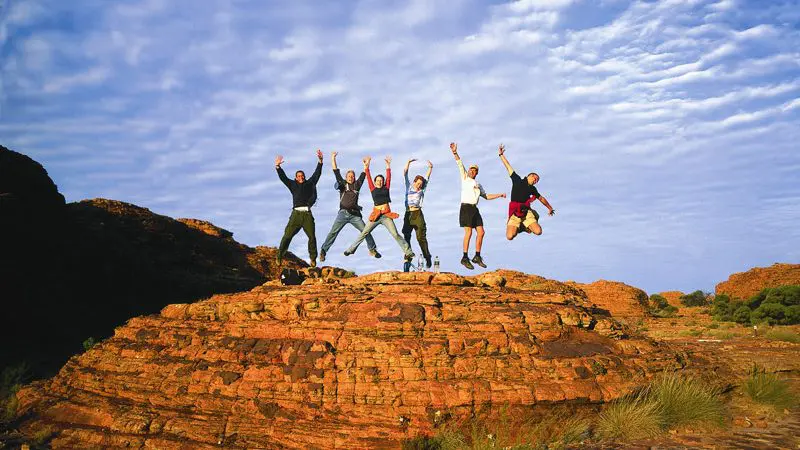 Group of six adventurers leaping on a scenic rocky hill, blue sky above, during a thrilling 3-Day Uluru Tour from Alice Springs.