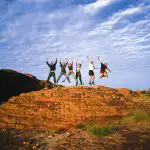 A group of six friends leap on a scenic rocky outcrop with vibrant blue skies during a 3-night Uluru adventure from Alice Springs.