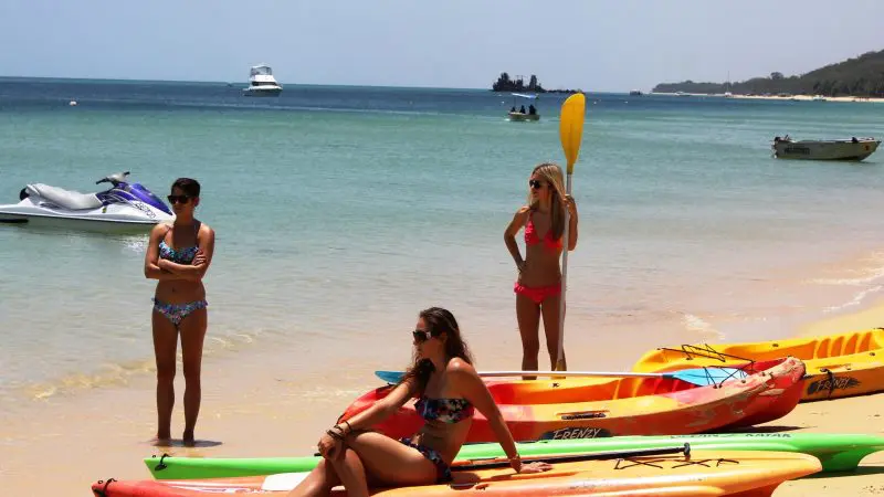 Three women in swimming costumes relax on kayaks at a sunny Moreton Island beach during a Wrecks Adventure Tour, enjoying the sea views.
