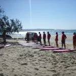 Surf students in matching red shirts stand on sunlit sand beside surfboards, preparing to surf Australia’s renowned longest wave.