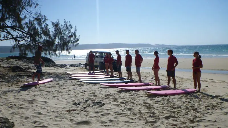 Surf students in matching red shirts stand on sunlit sand beside surfboards, preparing to surf Australia’s renowned longest wave.