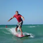 Man in bright red shirt skilfully surfs a small wave at Learn To Surf Australia's Longest Wave, clear blue sky overhead.