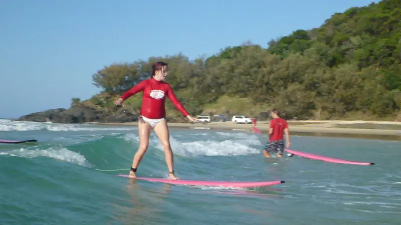 Surfer in a vibrant red shirt rides the waves near lush trees and parked cars at Learn To Surf Australia’s Longest Wave Beach Drive.