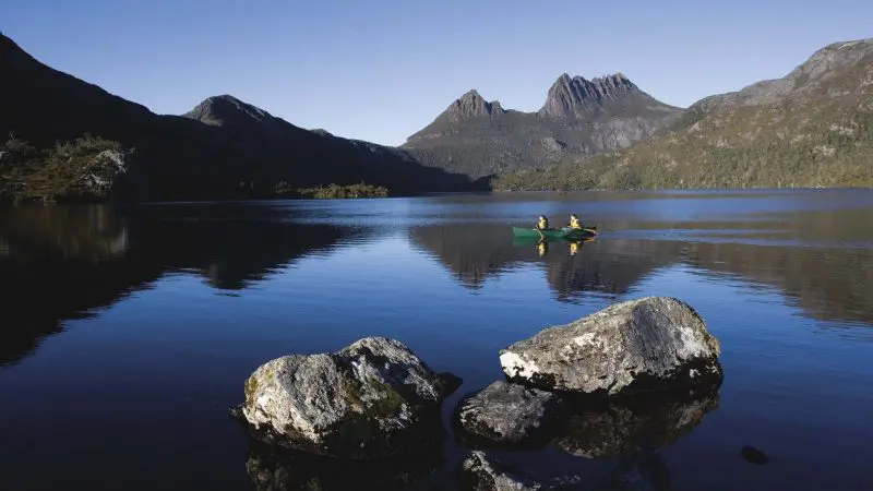Adventurers canoe on a serene Tasmanian lake surrounded by large rocks, showcasing the 6 Day Tasmania Explorer Tour experience.