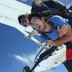 Two skydivers tandem jumping from an aeroplane over Sydney, one showing excitement and fear, freefalling against a clear blue sky.