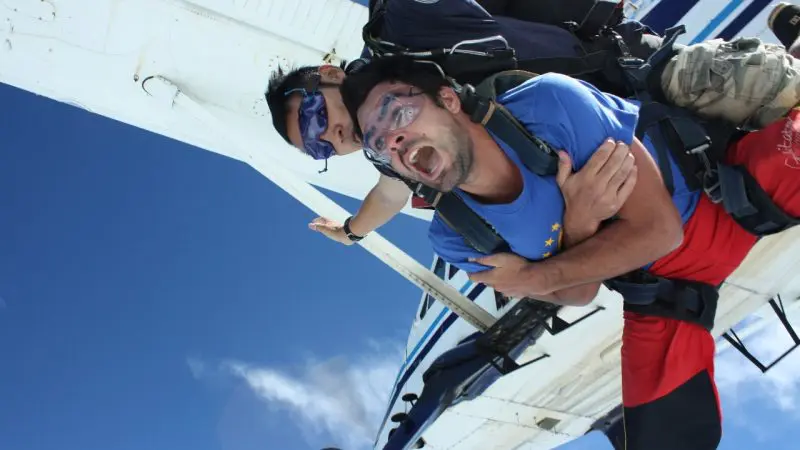 Two skydivers tandem jumping from an aeroplane over Sydney, one showing excitement and fear, freefalling against a clear blue sky.