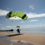 Skydiver with a vibrant green parachute from Sydney Tandem Skydive Extras lands on a stunning sandy ocean beach beneath clear blue skies.