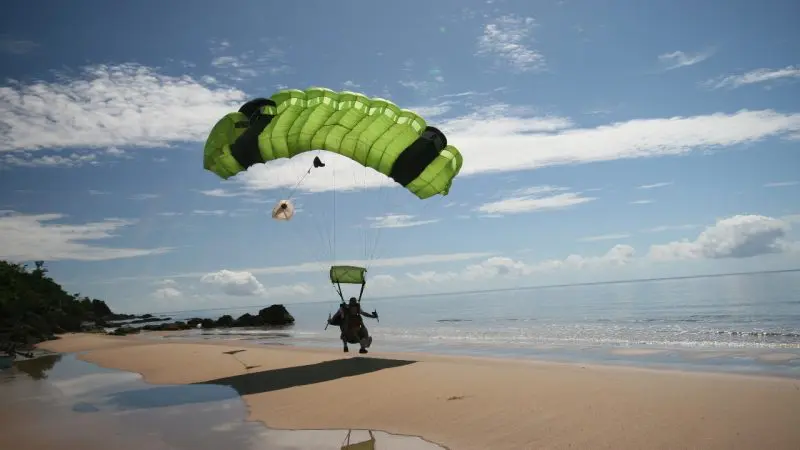 Skydiver with a vibrant green parachute from Sydney Tandem Skydive Extras lands on a stunning sandy ocean beach beneath clear blue skies.
