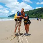 Two harnessed skydivers smile on Sydney’s sandy beach, celebrating their Tandem Skydive Extras beneath a bright blue sky.