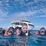 Three scuba divers signal OK underwater near a dive boat during a PADI Rescue Diver Course, demonstrating safe scuba diving skills.
