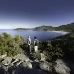 Two people stand on rugged rocks, taking in panoramic views of Wilsons Promontory Bay during a Go West Tours 1 Day guided tour.