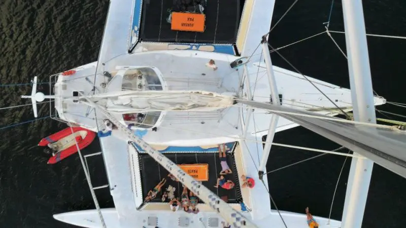 Stunning aerial view of Whitsundays catamaran sailing; people lounging on deck, red dinghy nearby, turquoise water surrounds.