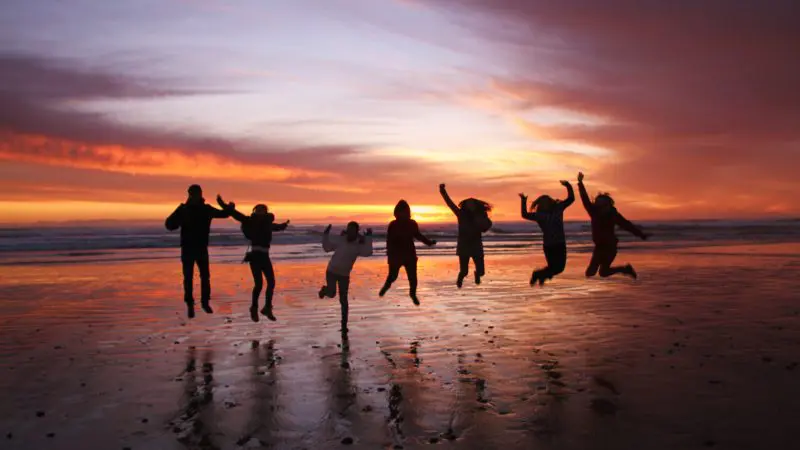 Seven people leaping with excitement on a beach at sunset during the 5 Day Famous 5 Tasmania Tour, showcasing travel joy and adventure.