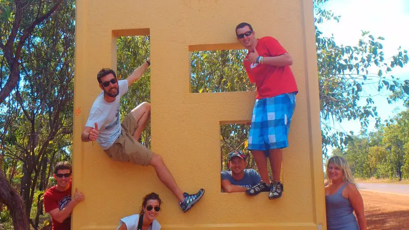 Six adventurers pose energetically by the iconic Kakadu National Park sign during a thrilling Kakadu Koolpin Tour camping experience.