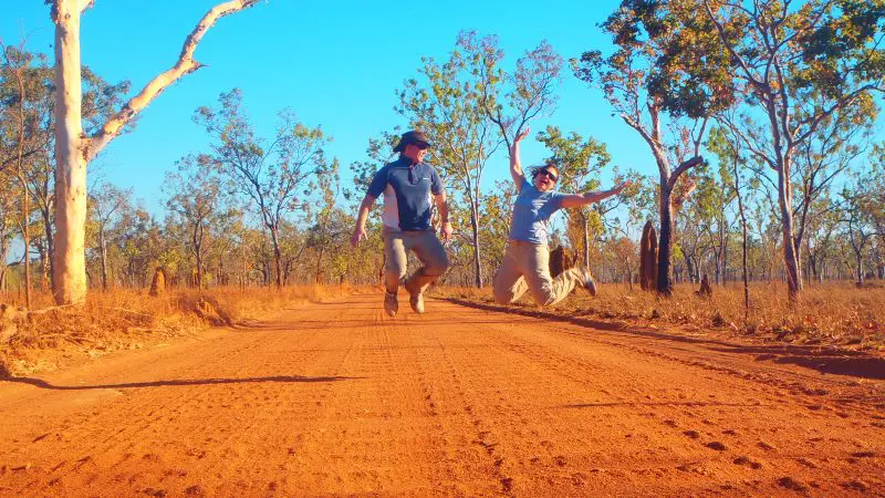 Two happy adventurers leap on a red dirt road in Kakadu National Park, enjoying 3 Day Camping Tour under clear blue skies, June–Sept.