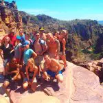 Tour group stands atop rocky cliffs on a Kakadu adventure, overlooking scenic canyon, winding river, lush trees, and bright blue sky.