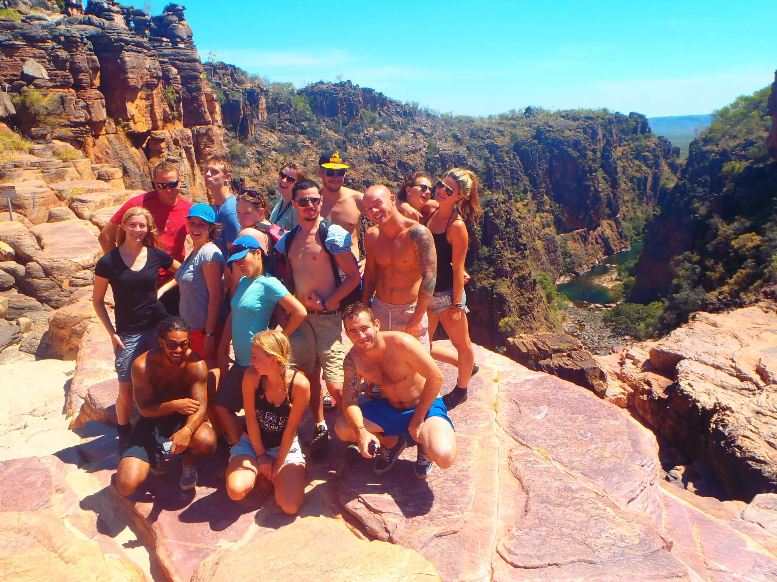 Tour group stands atop rocky cliffs on a Kakadu adventure, overlooking scenic canyon, winding river, lush trees, and bright blue sky.