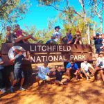 Tour group enjoying a 3-Day 4WD Kakadu Litchfield adventure, posing by the national park sign under sunny skies in lush woodland.