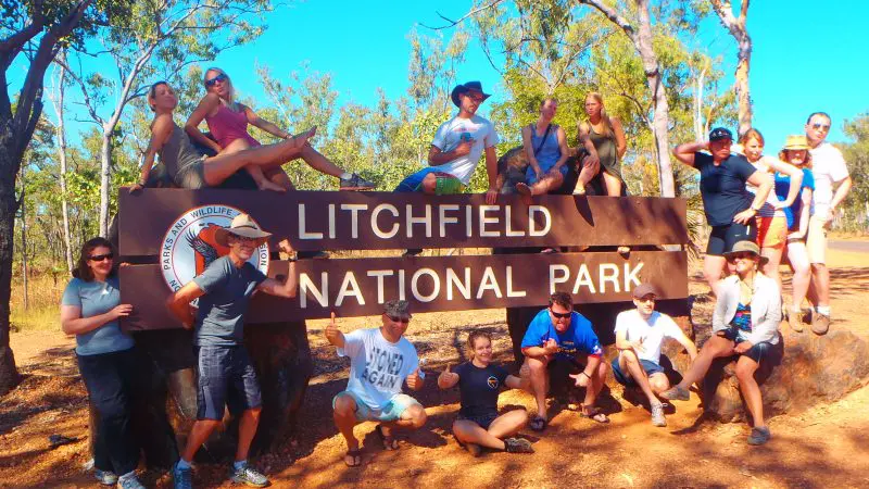 Tour group enjoying a 3-Day 4WD Kakadu Litchfield adventure, posing by the national park sign under sunny skies in lush woodland.