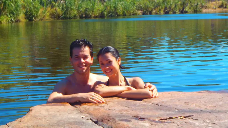 Happy couple swimming near a rock in crystal-clear blue lake, lush Kakadu Koolpin greenery all around, enjoying scenic Australia tour.